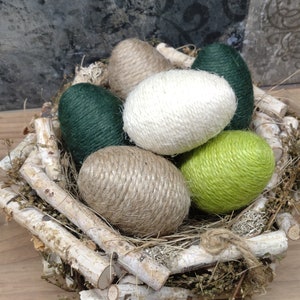 Könnte beinhalten: Ein Nest aus Zweigen und Moos beherbergt sechs dekorative Ostereier. Die Eier sind mit Schnur umwickelt und in den Farben Grün, Braun und Weiß gehalten.