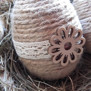 Op de afbeelding: Twee bruine paaseieren omwikkeld met touw en versierd met witte kant en houten bloemensnijwerk. De eieren liggen in een nest van droog gras.