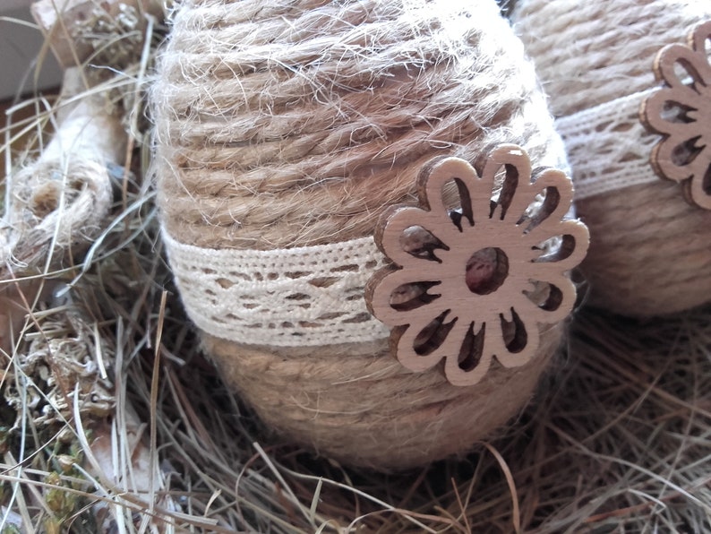 Op de afbeelding: Twee bruine paaseieren omwikkeld met touw en versierd met witte kant en houten bloemensnijwerk. De eieren liggen in een nest van droog gras.