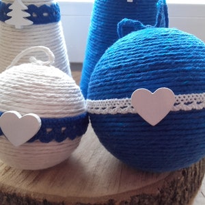 May include: Two blue and white yarn ornaments with white hearts and lace trim. The ornaments are sitting on a wooden surface.