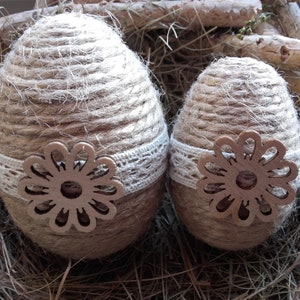 Puede incluir: Dos huevos de Pascua marrones envueltos en cuerda y decorados con encaje blanco y un recorte de flor de madera marrón.