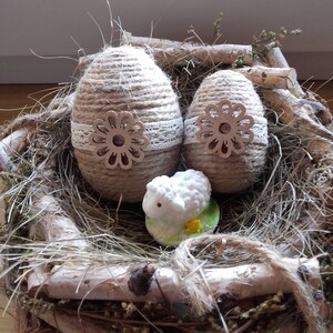 Op de afbeelding: Twee bruine, met touw omwikkelde paaseieren met witte kant en houten bloemaccenten, genesteld in een nest van takken en stro met een wit keramiek lammetjesfiguur.