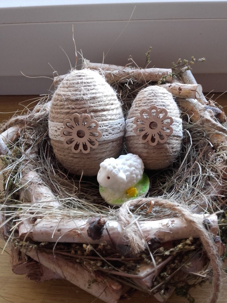 Op de afbeelding: Twee bruine, met touw omwikkelde paaseieren met witte kant en houten bloemaccenten, genesteld in een nest van takken en stro met een wit keramiek lammetjesfiguur.