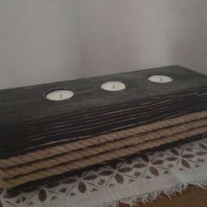 Puede incluir: Un portavelas de madera rústico con tres pozos para velas, decorado con cuerda natural. El portavelas está teñido de negro y tiene un acabado desgastado.
