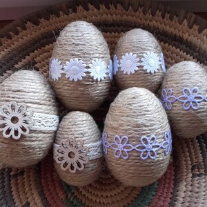 Op de afbeelding: Zes bruine paaseieren omwikkeld met touw en versierd met witte en paarse kantbloemen. De eieren liggen in een geweven mand.