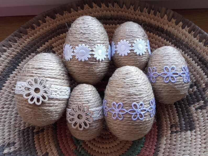Op de afbeelding: Zes bruine paaseieren omwikkeld met touw en versierd met witte en paarse kantbloemen. De eieren liggen in een geweven mand.