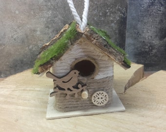 Könnte beinhalten: Ein kleines Vogelhaus aus Holz mit einem moosbewachsenen Dach, einer weißen und braunen Kordel-Außenhülle und einem Ausschnitt eines Vogels auf einem Ast. Das Vogelhaus hat einen weißen Sockel und eine weiße Schnur zum Aufhängen.