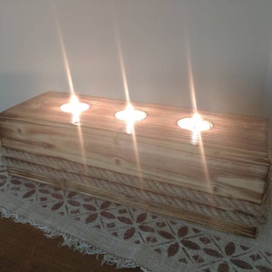 Puede incluir: Un portavelas de madera con tres huecos para velas. El portavelas está hecho de madera natural con un ribete de cuerda y tiene un aspecto rústico.