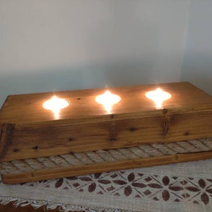 Puede incluir: Un portavelas de madera rústico con tres pozos para velas y un ribete de cuerda. El portavelas está hecho de madera recuperada y tiene un acabado natural.