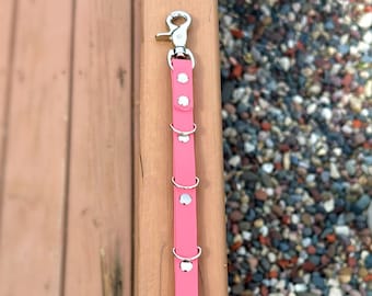 May include: A pink leather leash with silver metal rings and a silver metal clip. The leash is on a wooden surface.