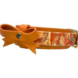 May include: Orange dog collar with a bow detail. The collar features a colorful abstract pattern in shades of orange, yellow, and white. The bow is a solid orange color, and the hardware is gold-toned.