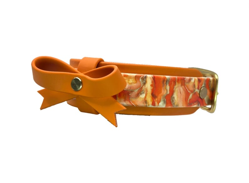 May include: Orange dog collar with a bow detail. The collar features a colorful abstract pattern in shades of orange, yellow, and white. The bow is a solid orange color, and the hardware is gold-toned.