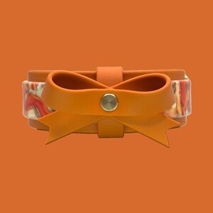 May include: An orange leather bracelet with a bow detail. The bow is made of the same orange leather and is centered on the bracelet. The bracelet has a patterned band with red, white, and orange colors. A silver button secures the bow.