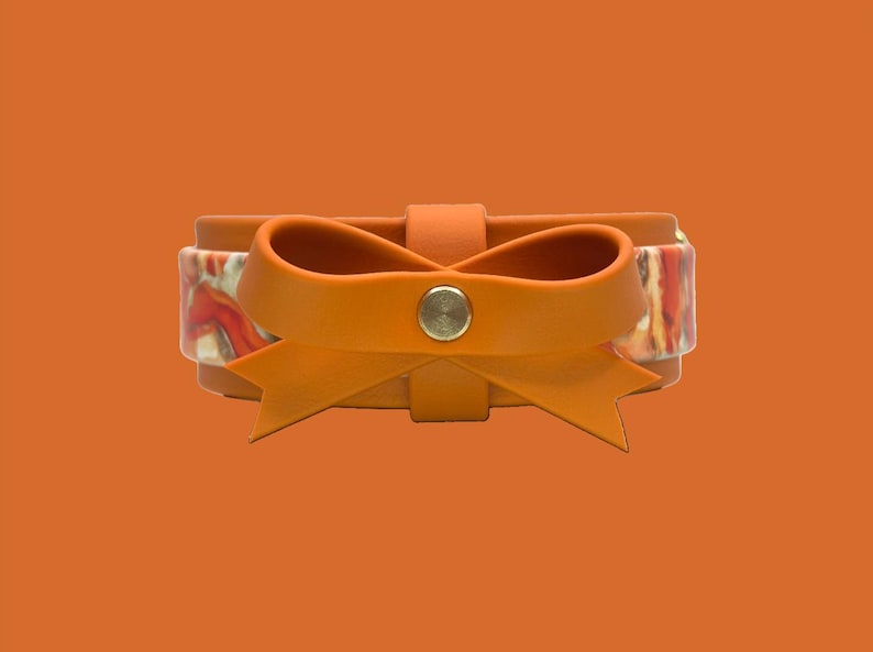 May include: An orange leather bracelet with a bow detail. The bow is made of the same orange leather and is centered on the bracelet. The bracelet has a patterned band with red, white, and orange colors. A silver button secures the bow.