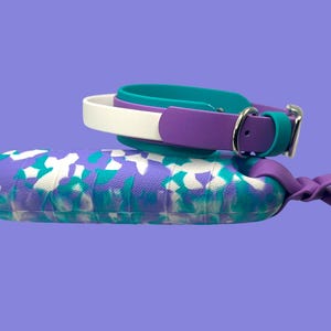 May include: A purple, teal, and white patterned pet carrier with matching collars. The carrier has a rounded shape and a knotted handle. The collars are teal, white, and purple with silver buckles. The background is a solid purple.