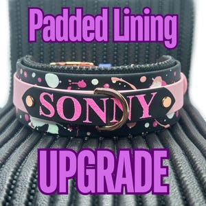 Op de afbeelding: Een zwart en roze hondenhalsband met witte verfspatten en de naam "SONNY" in roze letters. De halsband heeft een gevoerde voering en een gouden metalen D-ring.