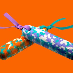 K&ouml;nnte beinhalten: Zwei bunte Springseile mit gemusterten Griffen. Eines hat ein orangefarbenes, t&uuml;rkisfarbenes und wei&szlig;es Muster, das andere ein lila, t&uuml;rkisfarbenes und wei&szlig;es Design. Beide haben farblich passende Griffe und Riemen vor einem orangefarbenen Hintergrund.