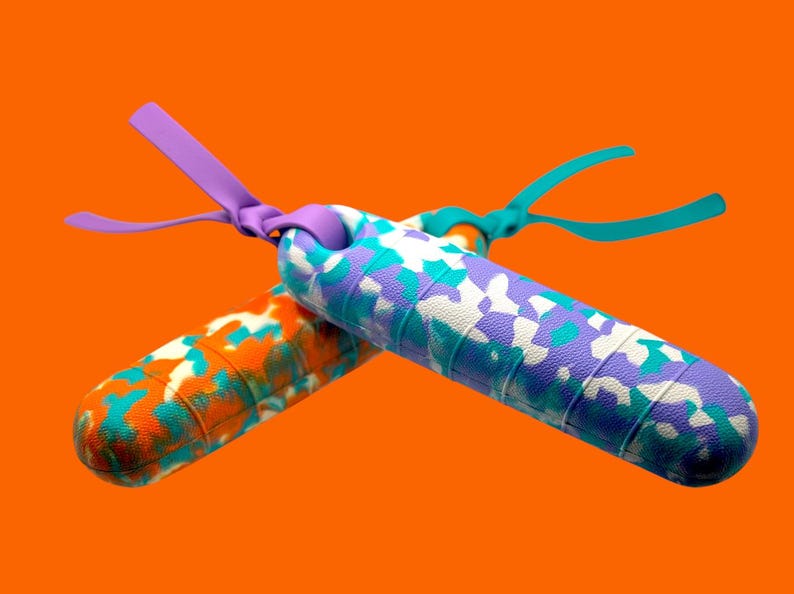 K&ouml;nnte beinhalten: Zwei bunte Springseile mit gemusterten Griffen. Eines hat ein orangefarbenes, t&uuml;rkisfarbenes und wei&szlig;es Muster, das andere ein lila, t&uuml;rkisfarbenes und wei&szlig;es Design. Beide haben farblich passende Griffe und Riemen vor einem orangefarbenen Hintergrund.