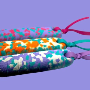 K&ouml;nnte beinhalten: Drei bunte Springseile mit gemusterten Griffen. Die Griffe weisen ein Camouflage-Design in Lila, Orange und Wei&szlig; mit Akzenten in Pink, T&uuml;rkis und Gr&uuml;n auf. Jedes Seil hat eine farblich passende Schlaufe.