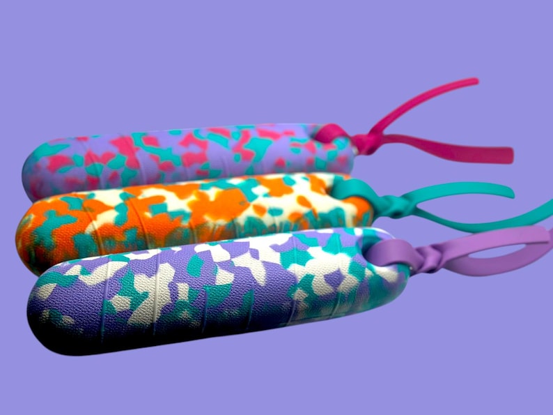 K&ouml;nnte beinhalten: Drei bunte Springseile mit gemusterten Griffen. Die Griffe weisen ein Camouflage-Design in Lila, Orange und Wei&szlig; mit Akzenten in Pink, T&uuml;rkis und Gr&uuml;n auf. Jedes Seil hat eine farblich passende Schlaufe.