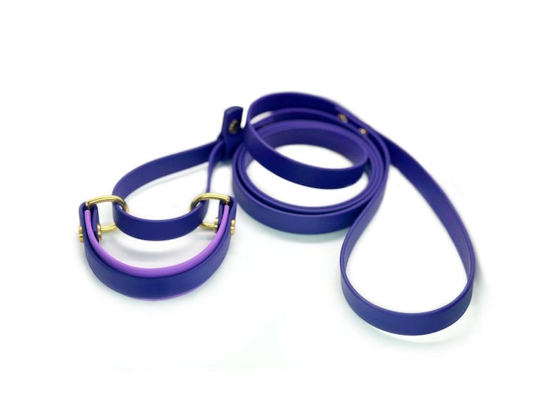 May include: A purple and lavender dog leash with a matching collar. The leash is made of a soft, flexible material and has a gold metal clasp.