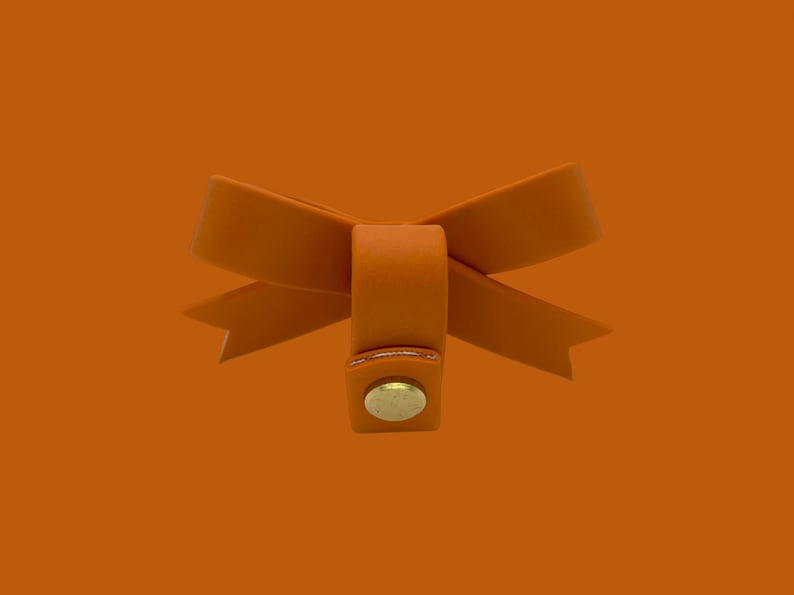 May include: An orange leather bow with a gold-colored button closure. The bow is centered against an orange background. The bow's design features two loops and two pointed ends.