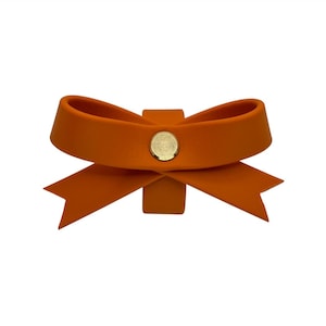 May include: An orange bow-shaped accessory with a gold-colored circular accent in the center. The bow has two loops and two downward-pointing tails. The accessory is set against a white background.