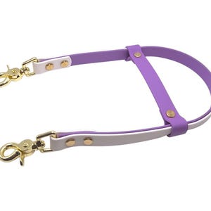 Op de afbeelding: Een tweekleurige paarse en witte riem met goudkleurige metalen hardware, inclusief twee draaibare karabijnhaken aan de uiteinden en ronde klinknagels die het gelaagde materiaal vastzetten, ontworpen met een schuiflus voor verstelbaarheid.