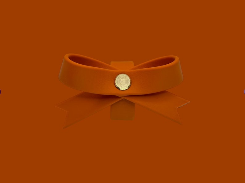 May include: Orange leather bow with a gold-colored button in the center. The bow is set against a matching orange background, creating a monochromatic effect. The bow's reflection is visible below.