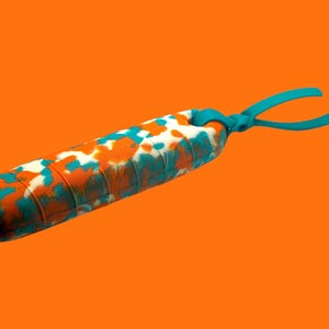 K&ouml;nnte beinhalten: Ein zylindrisches Massageger&auml;t mit strukturierter Oberfl&auml;che, das mit orangefarbenen, t&uuml;rkisfarbenen und wei&szlig;en Flecken gemustert ist. Es hat t&uuml;rkisfarbene Griffe und ist vor einem orangefarbenen Hintergrund platziert.