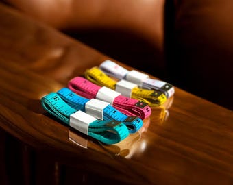 May include: Four colorful measuring tapes with white labels on a wooden surface. The tapes are blue, pink, yellow, and white.