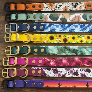 May include: A collection of colorful dog collars with various patterns, including floral, abstract, and sunflower designs. Each collar features a buckle and rivets, with colors like orange, green, blue, yellow, pink, and purple. The collars are displayed on a wooden surface.