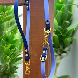 May include: Two dog leads, one blue and one purple, with gold hardware and a clip. The leads are hanging on a wooden post.