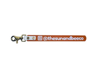 Könnte beinhalten: Ein lederner Schlüsselanhänger in Orange mit einem goldfarbenen Verschluss und weißen Lederakzenten. Der Riemen zeigt das Instagram-Logo und den Text "@thesunandbeeco" in Weiß.