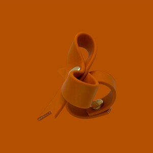 May include: An orange leather accessory with a gold-colored metal clasp. The leather is folded and looped, creating a unique shape. The background is a solid orange color.