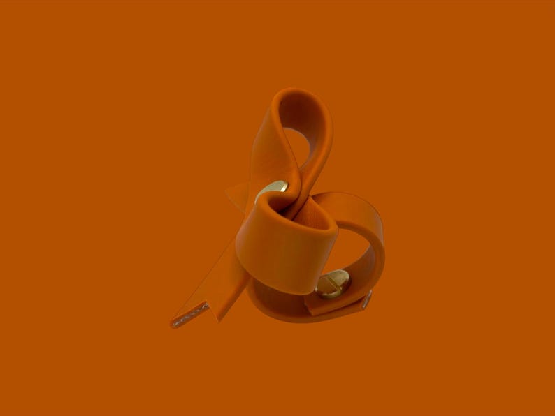 May include: An orange leather accessory with a gold-colored metal clasp. The leather is folded and looped, creating a unique shape. The background is a solid orange color.