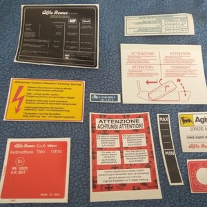 May include: A selection of vintage Alfa Romeo stickers and labels. The collection includes a black label with the Alfa Romeo logo and AGIP branding, warning labels, and technical specifications. Various sizes and shapes.