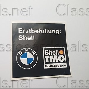 Puede incluir: Una pegatina cuadrada negra con el logotipo de BMW y la marca Shell. El texto "Erstbefüllung: Shell" se muestra encima de los logotipos. El logotipo de Shell incluye el texto "TMO" y "Das Öl der Besten."