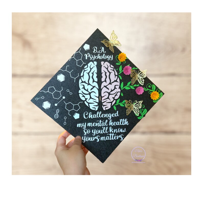 May include: Black graduation cap decorated with a brain, flowers, and butterflies. The text on the cap reads "B.A. Psychology Challenged my mental health so you'll know yours matters".