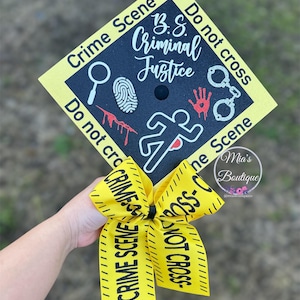 Op de afbeelding: Een gele afstudeerhoed met zwarte tekst die "Crime Scene B.S. Criminal Justice Do not cross" luidt en een zwart-witte illustratie van een vingerafdruk, een vergrootglas, bloederige voetafdrukken, een handafdruk en handboeien. De hoed heeft een geel lint met zwarte tekst die "Crime Scene Do Not Cross" luidt.