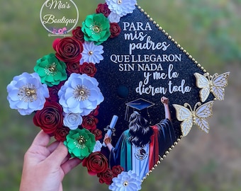 Puede incluir: Un birrete negro decorado con flores de papel rojas, verdes y blancas, y detalles de mariposas doradas. El birrete presenta texto en español y una ilustración de una graduada.