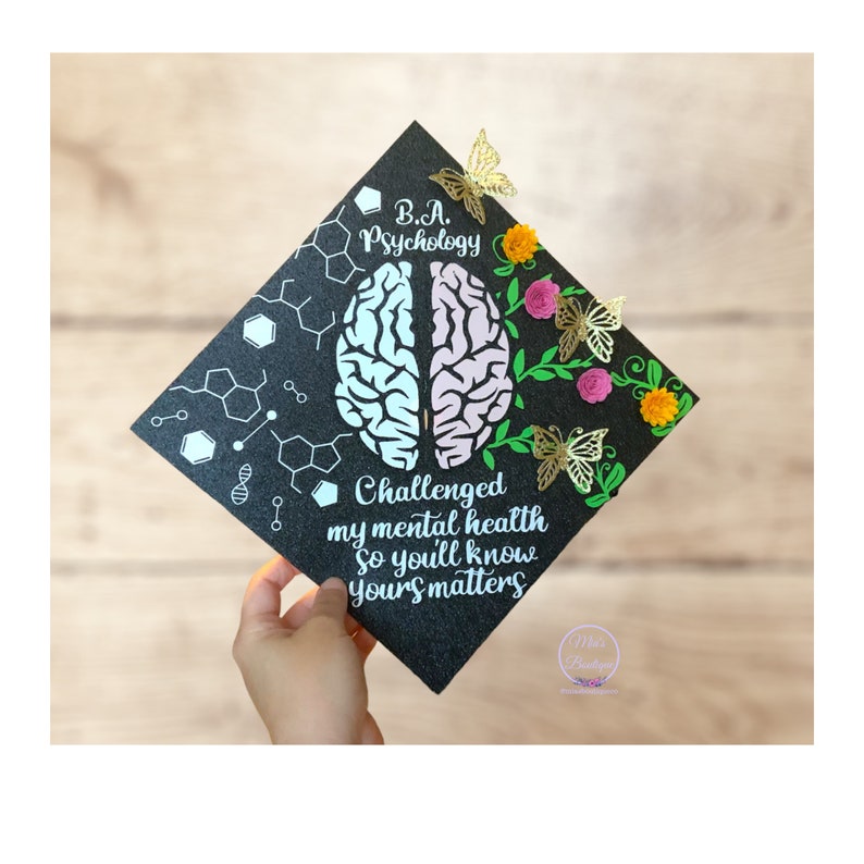 May include: Black graduation cap decorated with a brain, flowers, and butterflies. The text on the cap reads "B.A. Psychology Challenged my mental health so you'll know yours matters".