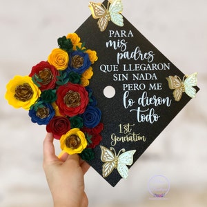May include: A black graduation cap decorated with paper flowers in red, yellow, blue, and green. The cap has gold glitter and the text "PARA mis padres QUE LLEGARON SIN NADA PERO ME lo dieron todo 1st Generation" in white.