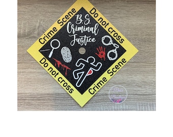 Puede incluir: Un gorro de graduación amarillo con cinta de escena del crimen en blanco y negro y una silueta negra de una persona corriendo. El texto en la cinta dice "Crime Scene Do Not Cross" y "B.S. Criminal Justice".
