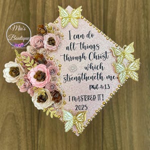 以下が含まれることがあります： 紙の花、蝶、真珠で飾られたピンク色の卒業キャップ。キャップには「I can do all things through Christ which strengtheneth me. Phil 4:13」と「I MASTERED IT! 2025」の文字が書かれています。