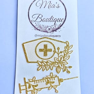 Puede incluir: Sombrero de enfermera y jeringa de purpurina dorada con detalles florales. El sombrero tiene un fondo blanco con una cruz dorada. La jeringa tiene un fondo blanco con una aguja dorada. Los detalles florales son de purpurina dorada y tienen un fondo blanco. El texto "Mia's Boutique" y "@miasboutiqueco" está impreso en la tarjeta.