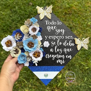 May include: A black graduation cap decorated with white paper flowers, gold glitter, and two gold butterflies. The cap has a blue and white striped band with the flag of El Salvador. The text on the cap reads "Todo lo que soy y espero ser, se lo debo a los que creyeron en mi".