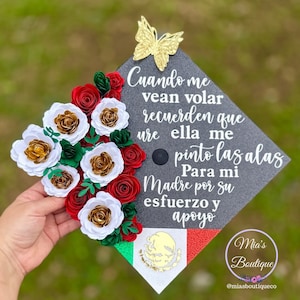 May include: Graduation cap decorated with red, white and gold paper flowers, a gold butterfly and a Mexican flag. The cap has a message in Spanish that reads: "Cuando me vean volar recuerden que ure ella me pinto las alas Para mi Madre por su esfuerzo y apoyo".