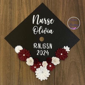 May include: Black graduation cap decorated with white and burgundy paper flowers. The cap has the text "Nurse Olivia RN, BSN 2024" written in white.