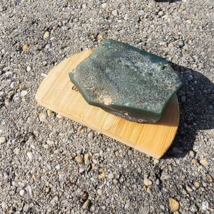 May include: A rough, dark green mineral specimen with a textured, uneven surface resting on a semi-circular, light-colored bamboo wood base. The mineral displays natural variations in color and surface depth, while the wooden base features a smooth, grain-patterned finish with a partial, partially visible dark text imprint on its surface.